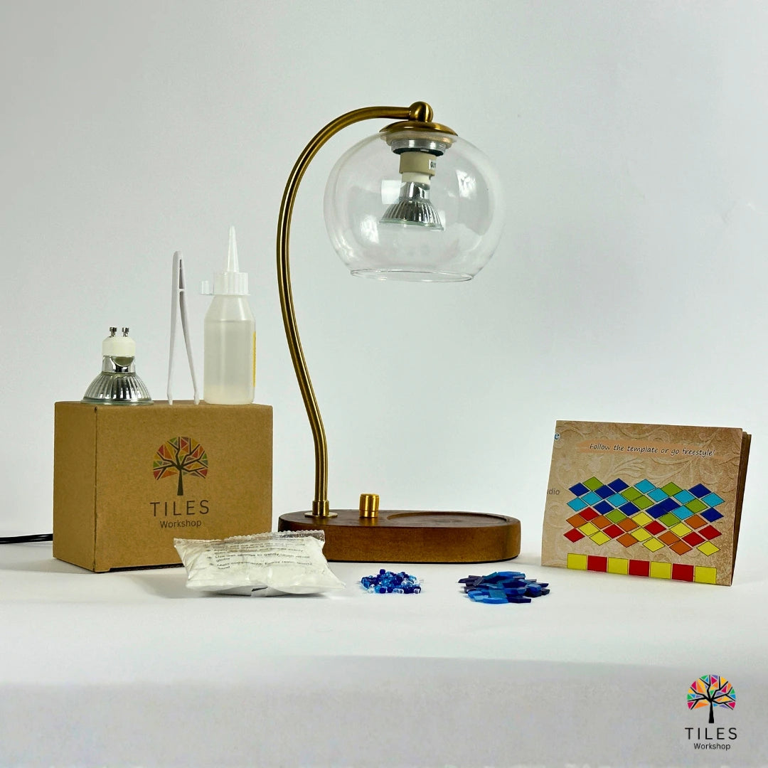 A mosaic tile kit by Tiles Workshop displayed on a white surface against a white background. The kit includes a brass gooseneck table lamp with a clear glass globe shade, a brown cardboard box, a small bottle, a small metal container, loose blue mosaic tiles, a small instruction card, and a finished sample mosaic tile panel.