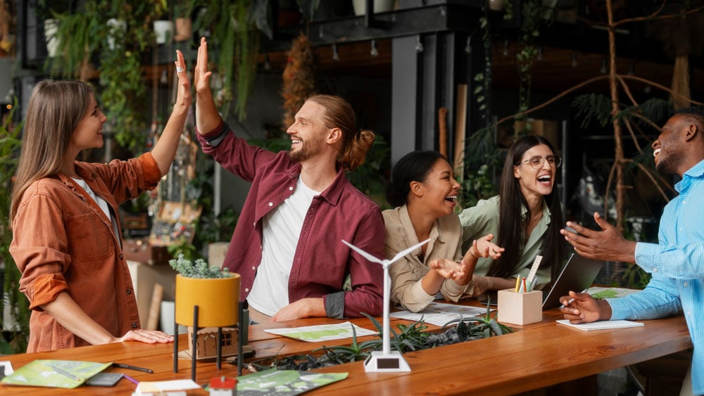 Five adults laugh and high-five at a wooden table during fun group activities.